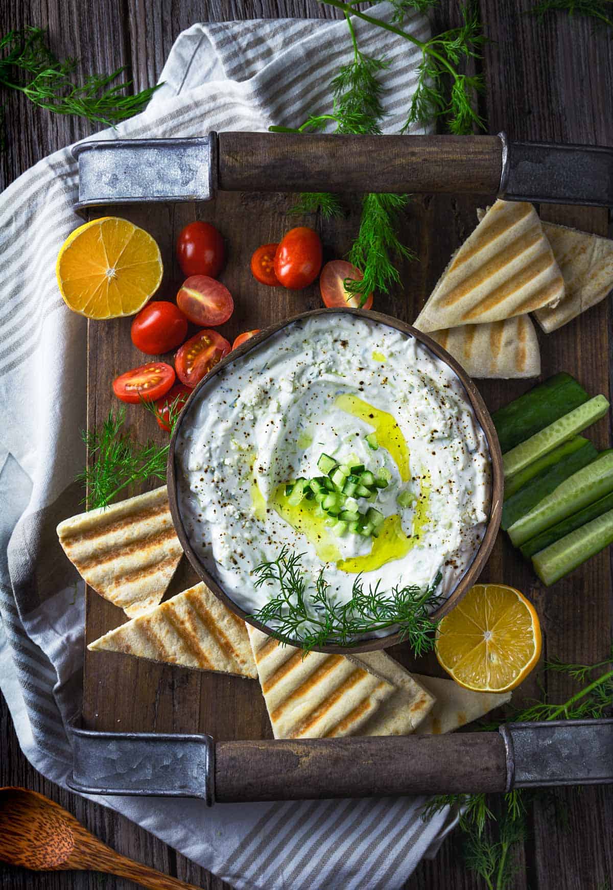 An overhead photo of yogurt dip with cucumber, dill, lemon, and garlic, served with vegetables and pita bread. 