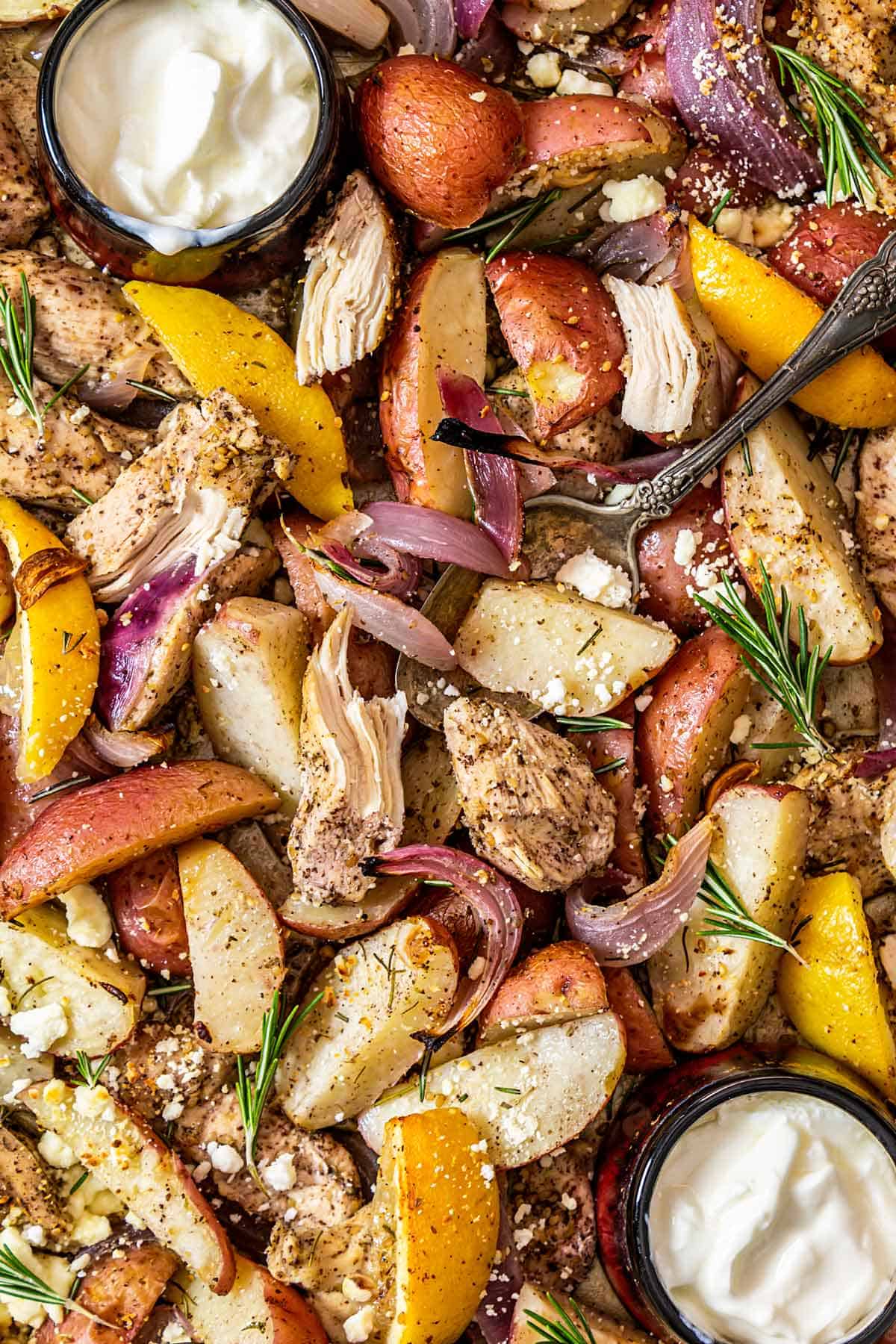 A close up photo of roasted chicken strips, potatoes, onions, lemon wedges, and two dishes with yogurt.