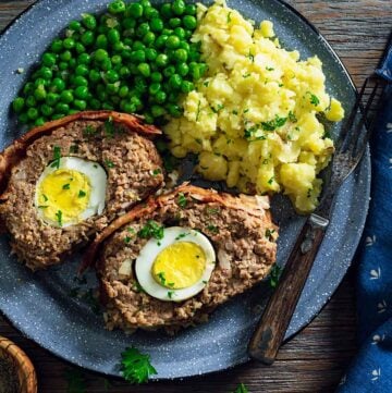 An overhead photo of two meatloaf slices showing the boiled eggs inside on a plate with mashed potatoes and green peas.