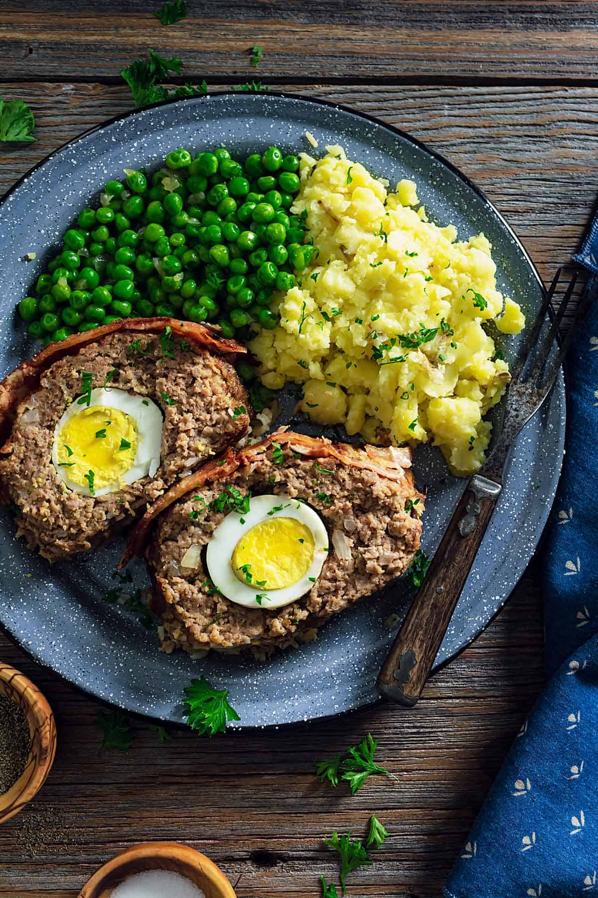 An overhead photo of two meatloaf slices showing the boiled eggs inside on a plate with mashed potatoes and green peas.