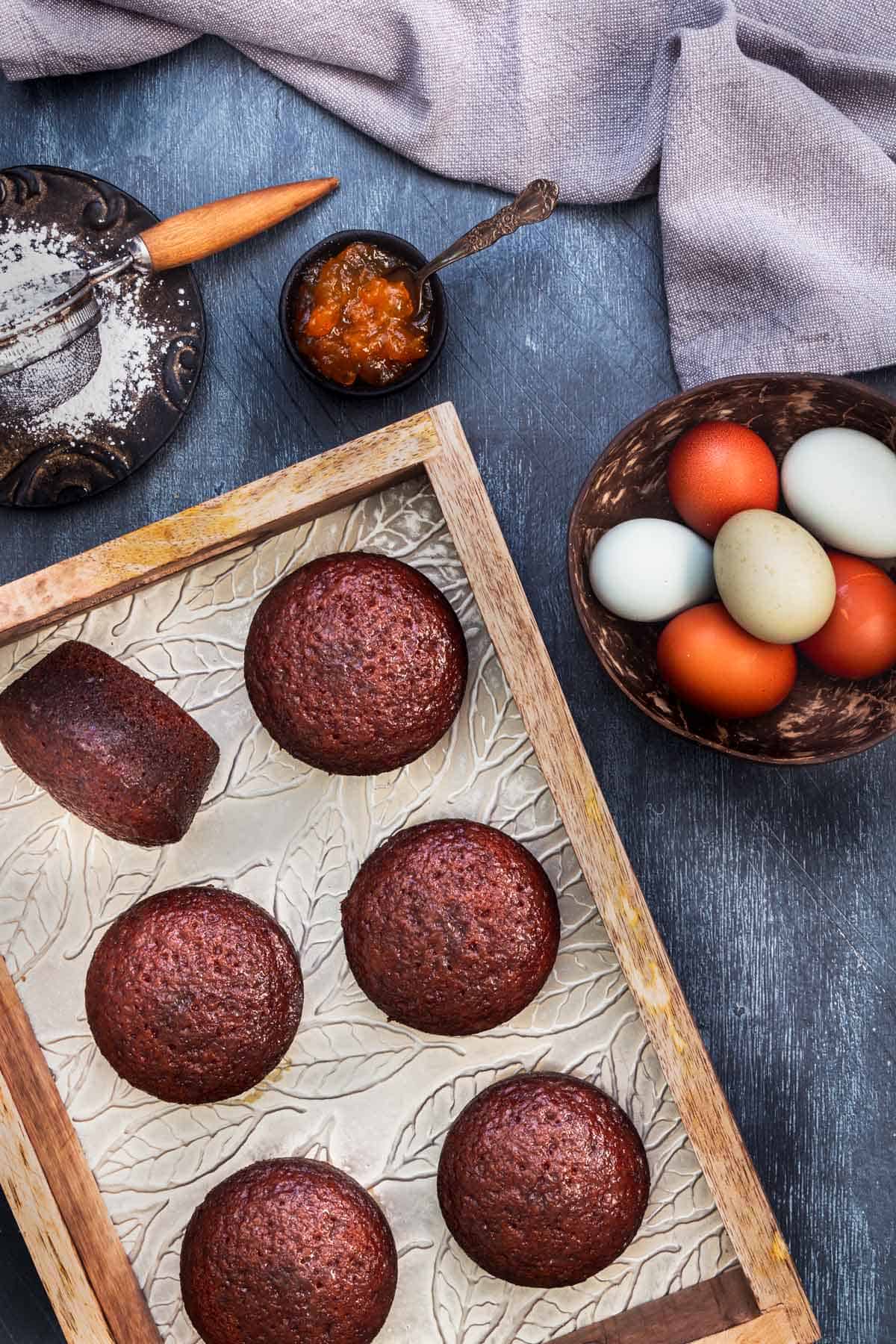 South African malva pudding cakes in a wooden tray on the table with eggs, jam, and powder sugar.