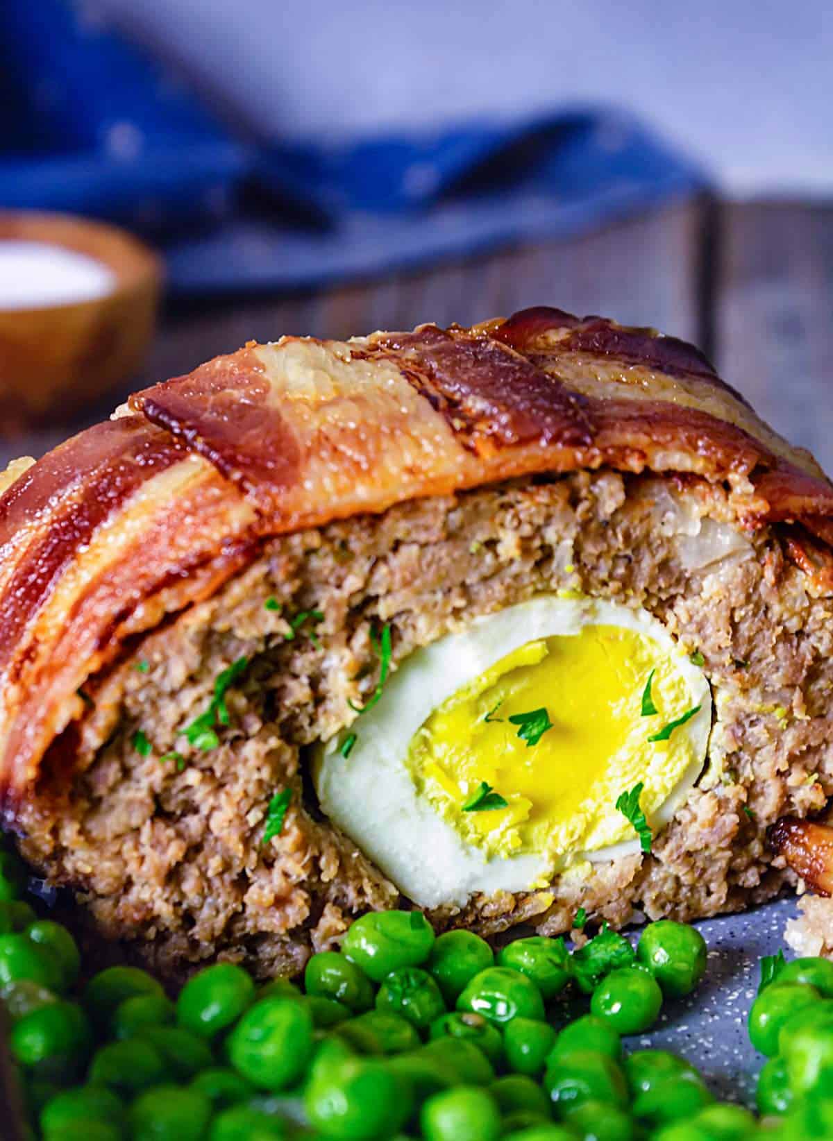 A close up of the sliced meatloaf with bacon on top and cooked egg in the middle with a side of peas.