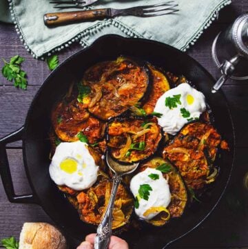 A person serving roasted eggplant with yogurt sauce from a skillet with a spoon.