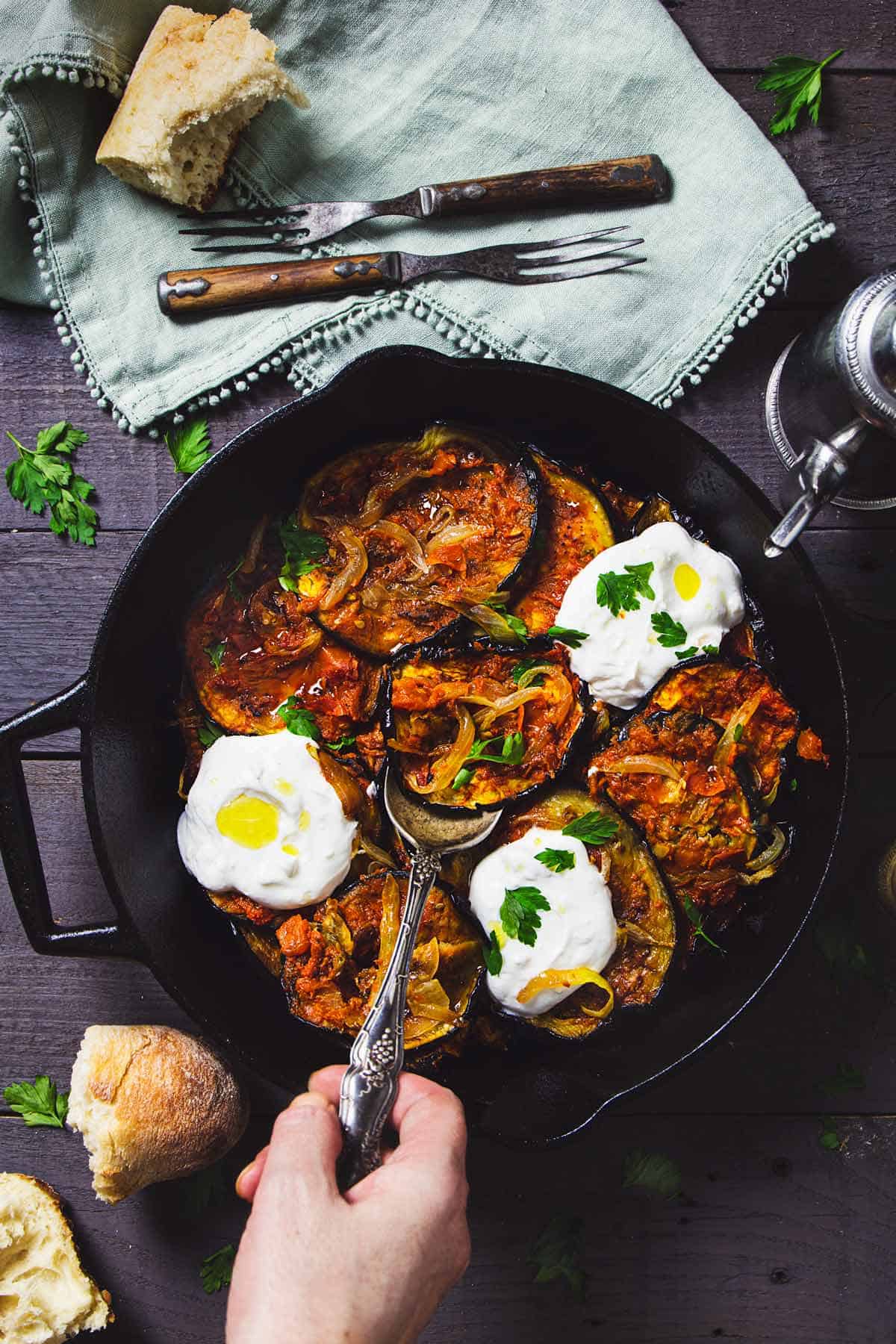 A person serving roasted eggplant with yogurt sauce from a skillet with a spoon.