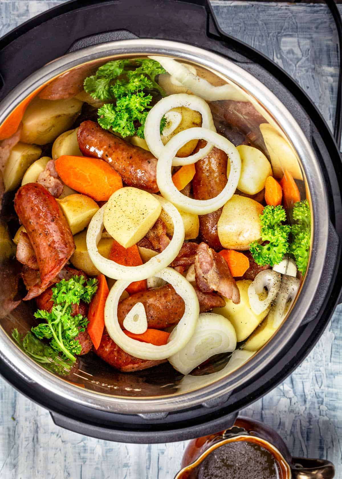 Potatoes, garlic, onions, carrots, and parsley added to the sausages and bacon in an Instant Pot.