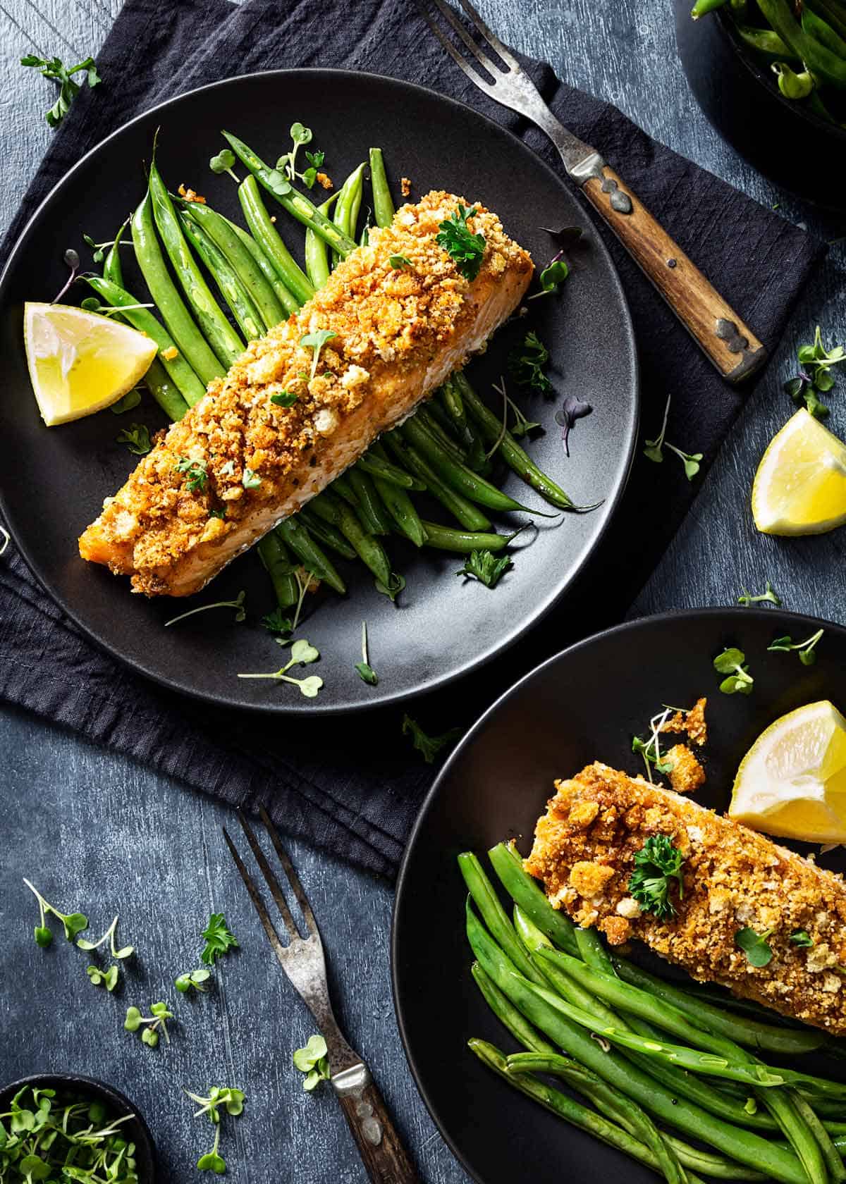 Two plated salmon filets over green beans with lemon wedges on a table.
