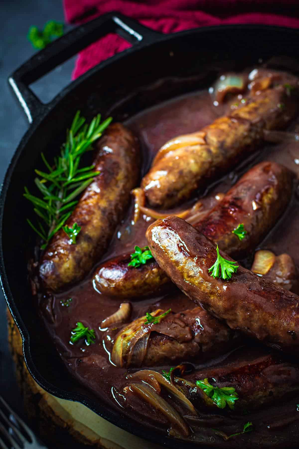 A close up of sausage bangers in a cast-iron skilled drenched in onion gravy and sprinkled with parsley.