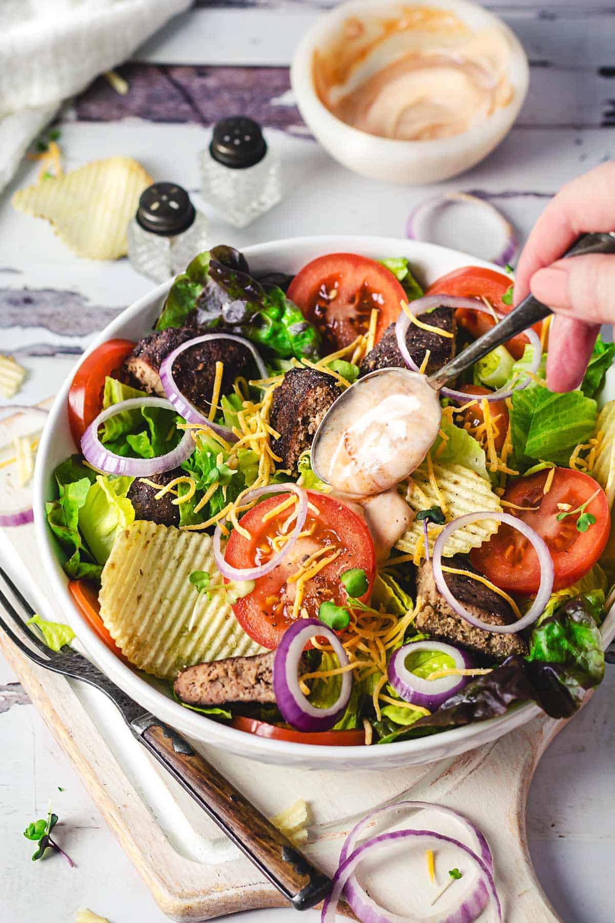 Adding big mac dressing over a cheeseburger salad in a bowl with a spoon. 