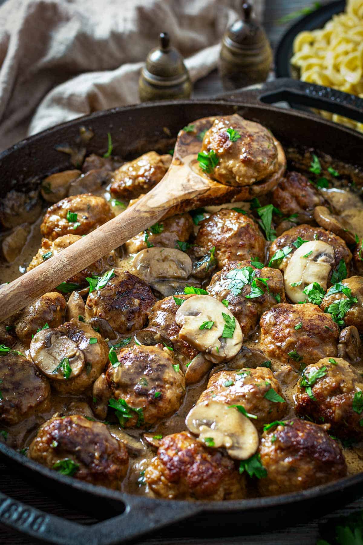 Meatballs in a cast-iron skillet with mushroom sauce and a wooden spoon.