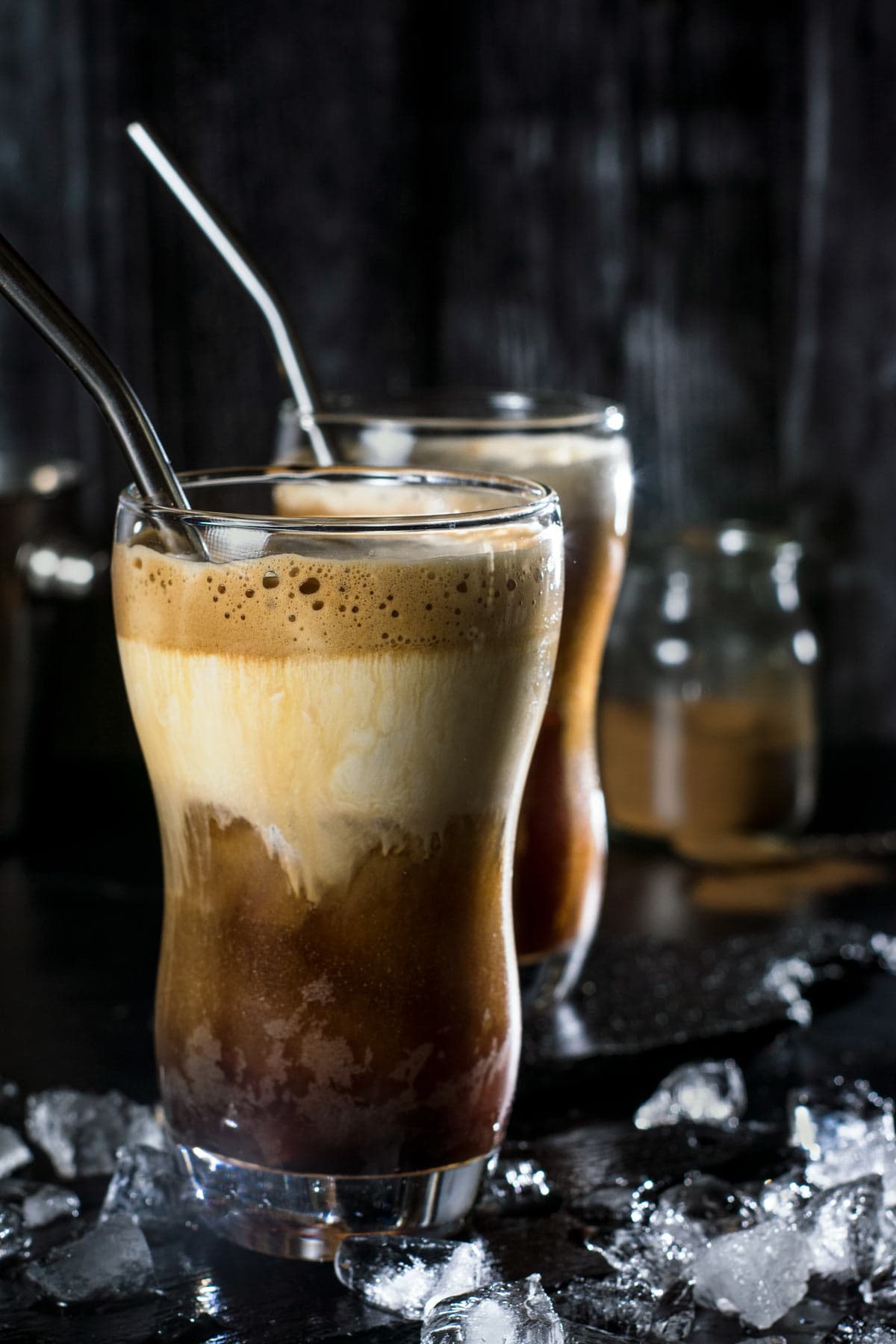 Two glasses of Greek frappe with metal straws on a black background with ice cubes.