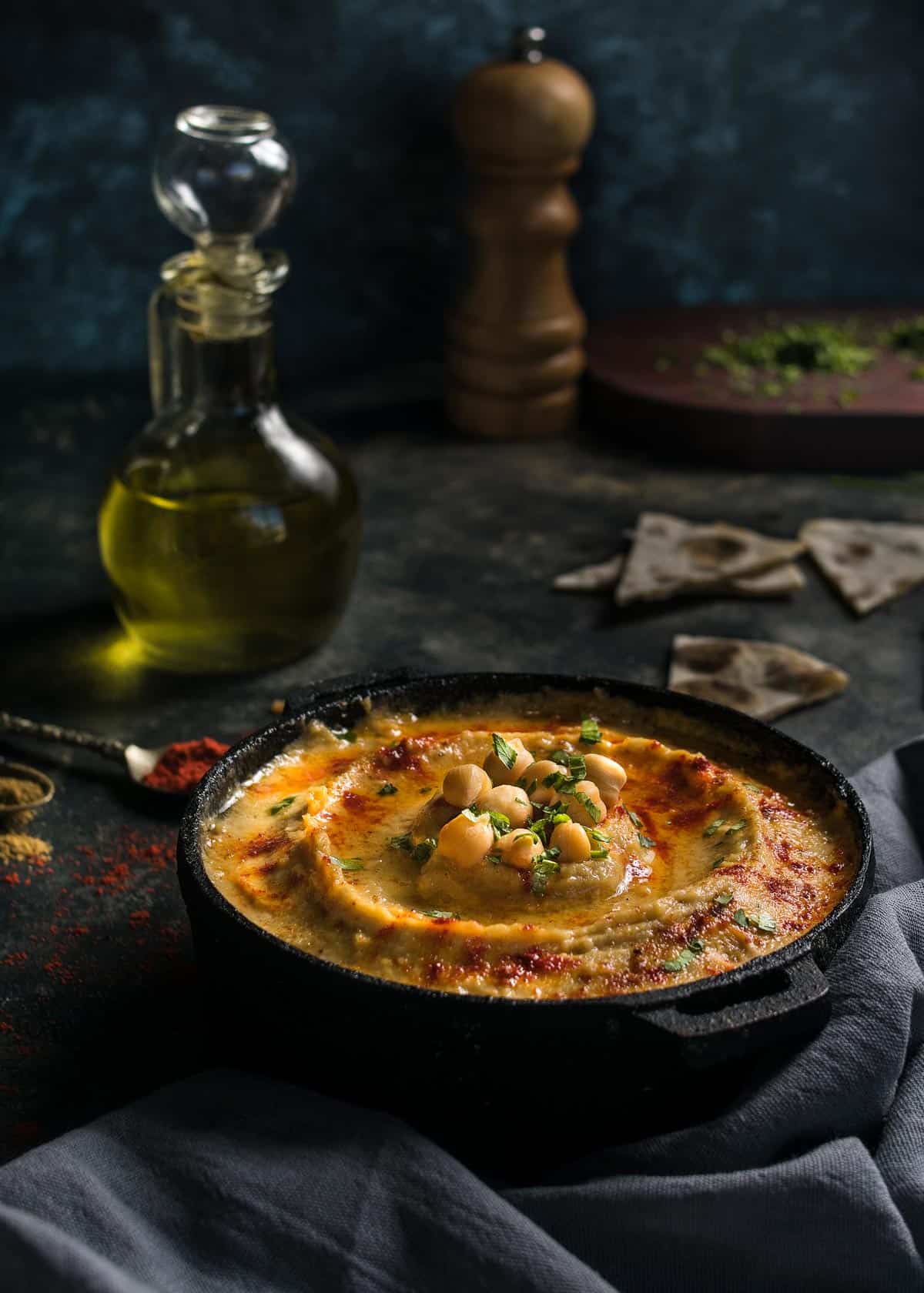 A dish with garlic hummus on a table with toasted pita bread triangles, olive oil, and pepper grinder.