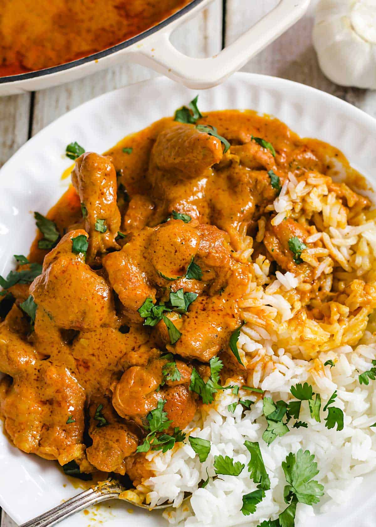 A close up of chicken tikka masala in a bowl next to white rice and garnished with cilantro leaves.