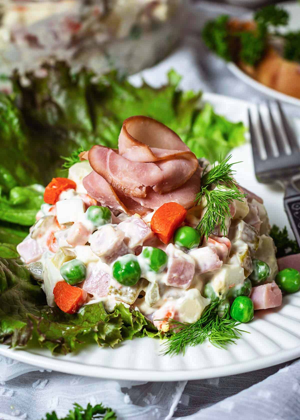 A close up serving of Russian potato salad with peas, carrots, eggs, and ham.